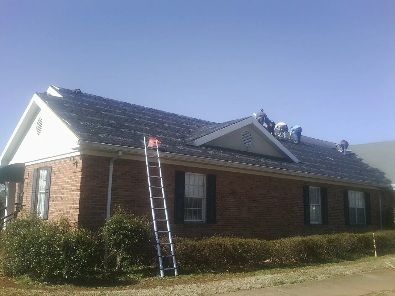 Crew tearing shingles off a brick ranch roof with a ladder set — Metal Roof Repair work by our crew in Polk City