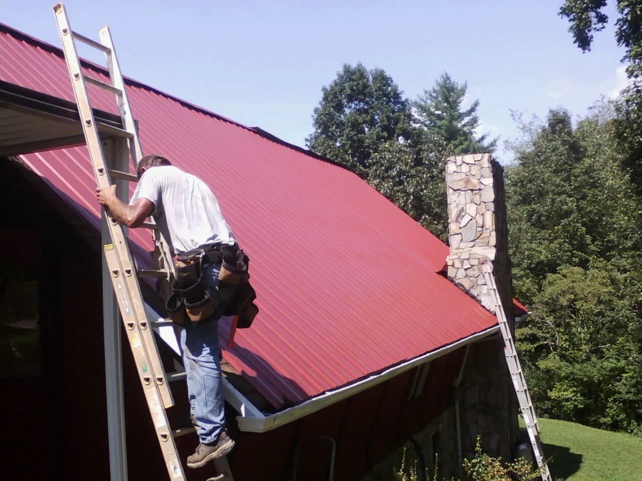 Roofer on a ladder beside a red metal roof and stone chimney — Soffit & Fascia Repair by Roofing Brothers in Polk City